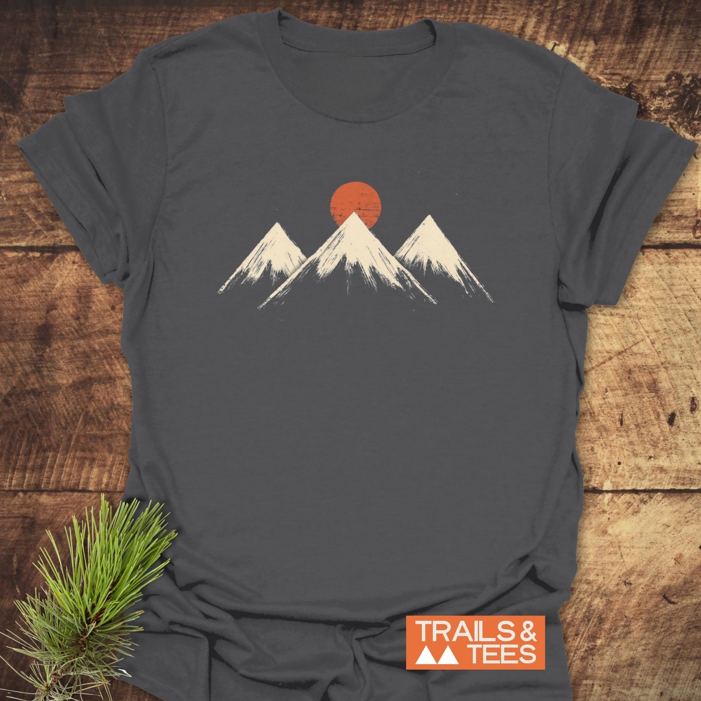 Three Mountains And Sun T-Shirt on wood, featuring a distressed mountain graphic with an orange sun, Trails & Tees logo, emphasizing adventure and comfort.