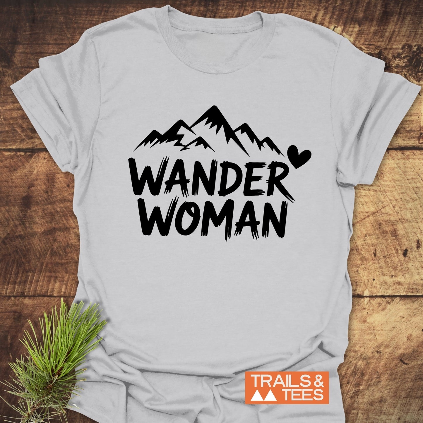 Wander Woman T-Shirt with black mountain peaks and text, laid on rustic wood. Crafted from soft cotton, designed for outdoor enthusiasts. Pine sprig styling accent.