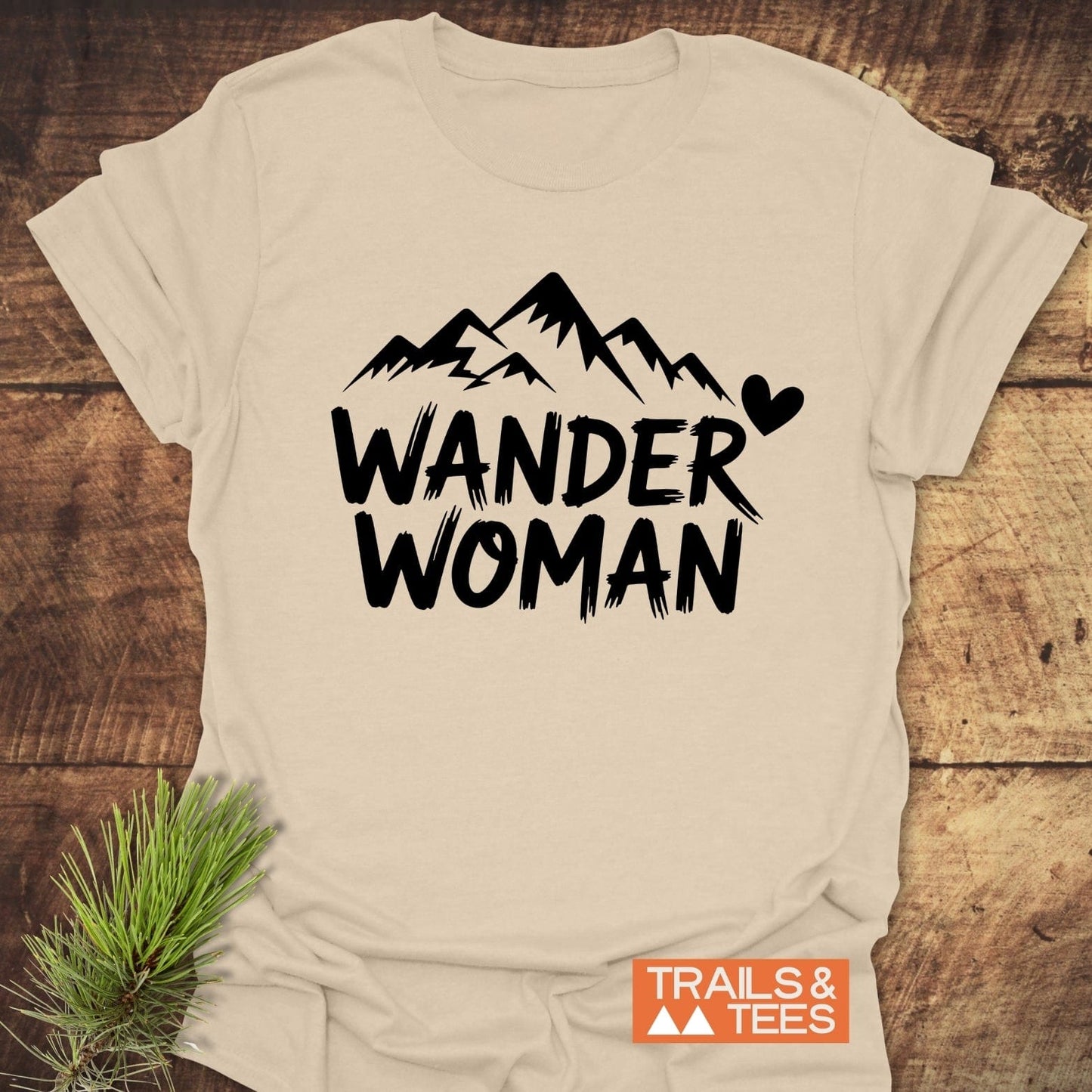Wander Woman T-Shirt featuring bold mountain graphic and text, crafted from soft cotton for comfort, with durable prints, laid flat with pine sprig accent.