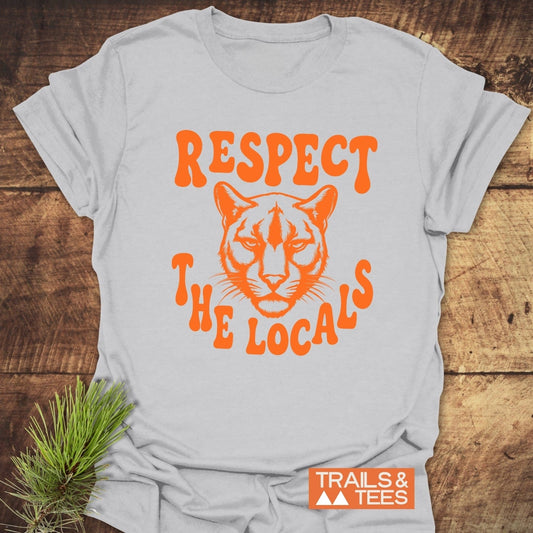 Respect The Locals, Cougar T-Shirt: Light-gray tee with bold orange cougar graphic, Respect The Locals text, and Trails & Tees logo, laid on wood.