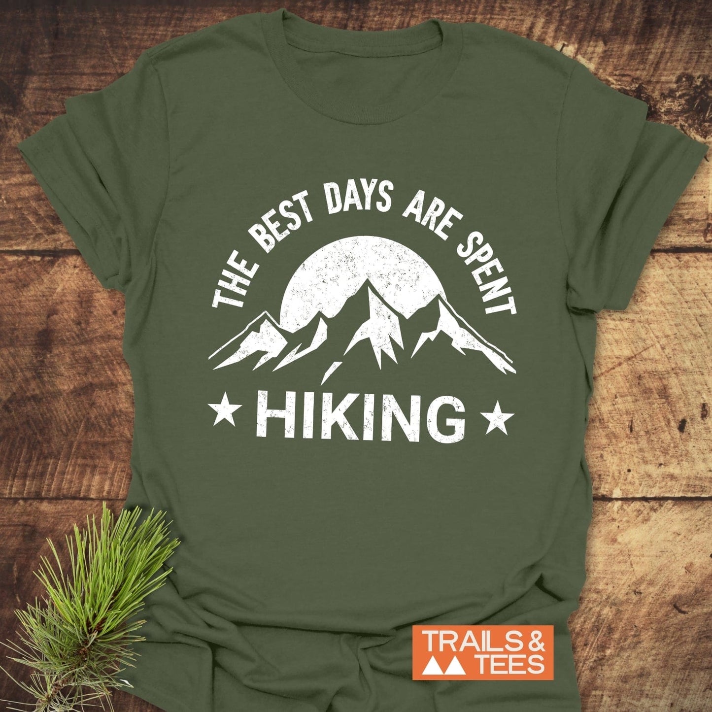 Olive-green t-shirt with a mountain and sun graphic, reading “The Best Days Are Spent Hiking,” beside a pine sprig and Trails & Tees logo.