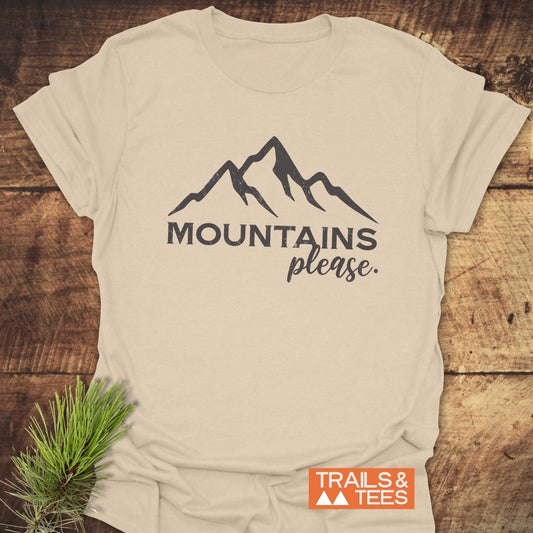 Mountains Please T-Shirt featuring a stylized mountain graphic and MOUNTAINS please text, with a Trails & Tees logo, on a rustic wooden background.