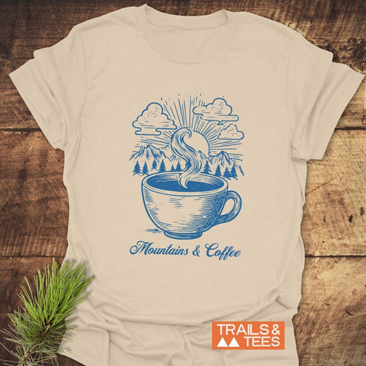 Mountains And Coffee T-Shirt featuring a blue coffee cup and mountain design, laid flat on wood. Includes TRAILS & TEES logo.