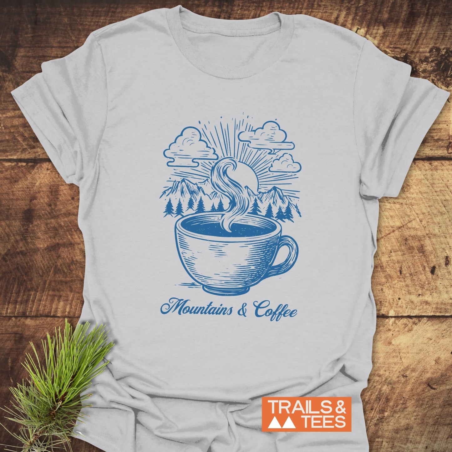 Mountains And Coffee T-Shirt featuring a blue steaming coffee cup with mountains, pine trees, and sun graphic. Part of the Trails & Tees collection.