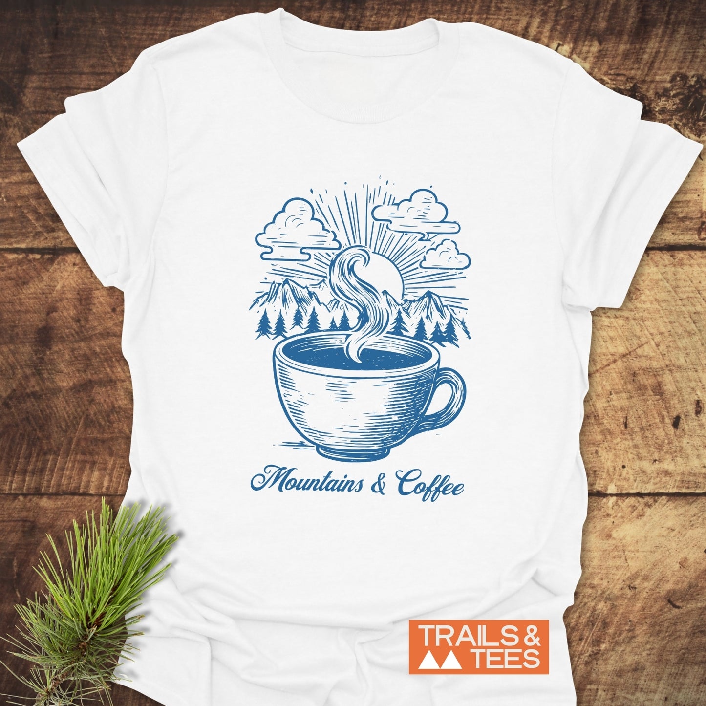 Mountains And Coffee T-Shirt featuring a blue coffee cup and mountain scene design, styled with an evergreen sprig, embodies outdoor adventure and comfort.