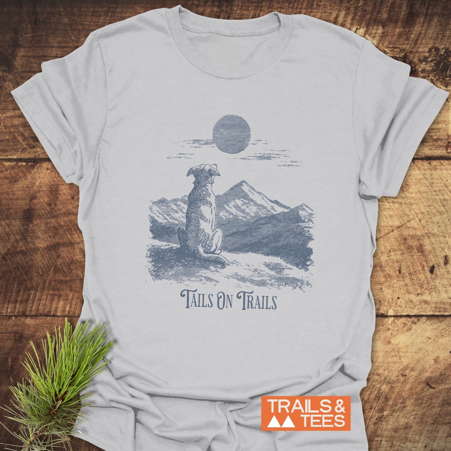 Tails On Trails T-Shirt featuring a dog and mountain print, laid on wood, symbolizing outdoor adventure. Made from soft cotton, pre-shrunk, durable prints.