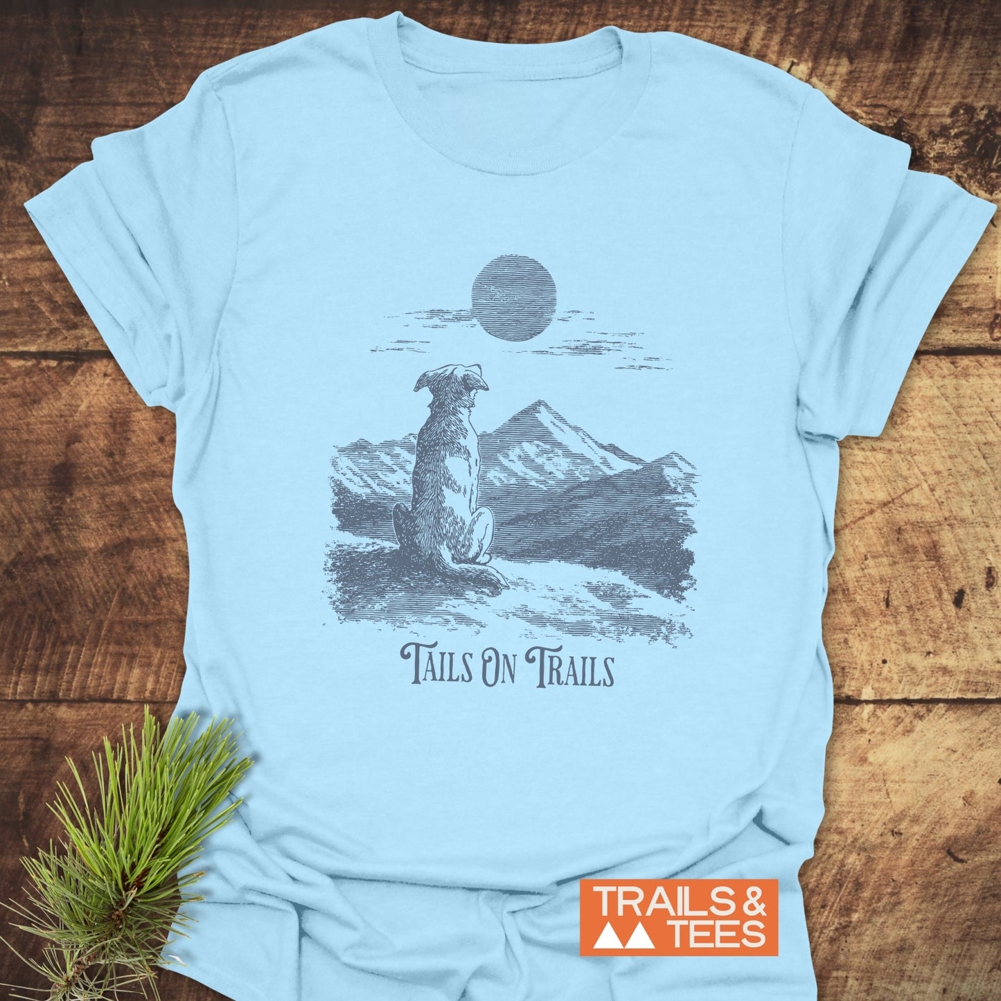 Light-blue Tails On Trails T-Shirt featuring a dog illustration with mountain scenery, crafted from soft cotton, highlighting comfort and durability for outdoor enthusiasts.