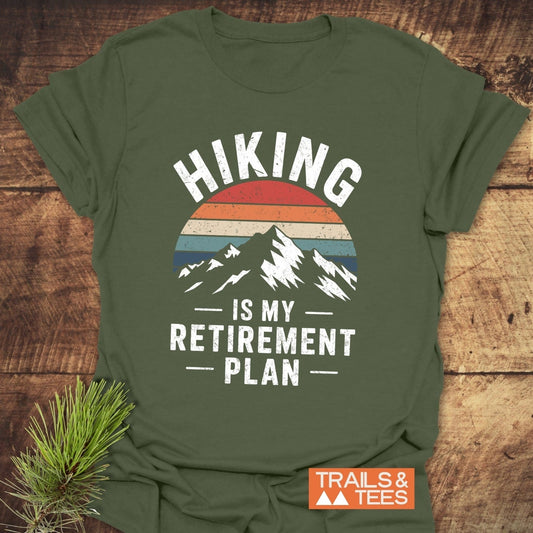 Olive-green T-shirt with mountain silhouette and sunset motif, featuring the slogan “Hiking Is My Retirement Plan,” beside a pine sprig and Trails & Tees logo.