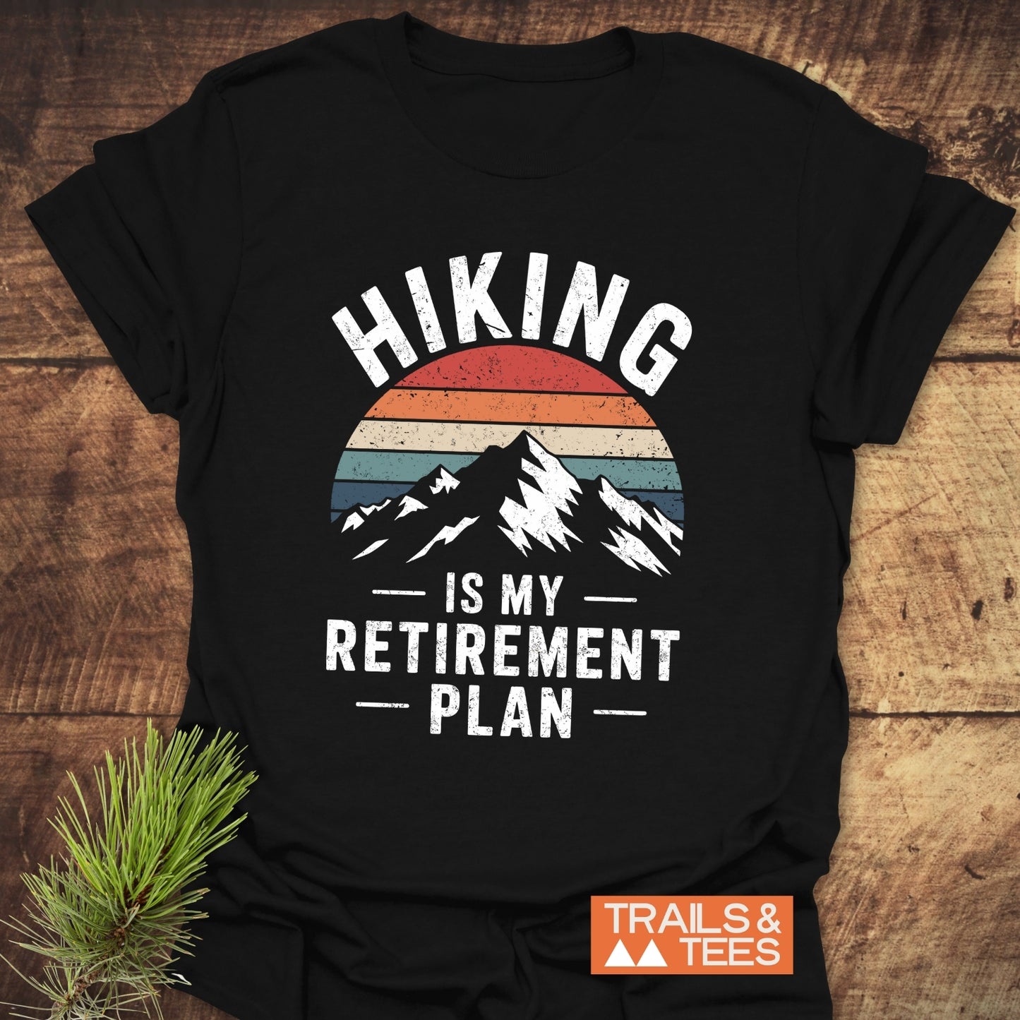 Hiking Is My Retirement Plan T-Shirt featuring a mountain graphic and slogan, laid on wood with pine branch, showcasing outdoor-ready, comfortable ring-spun cotton fabric.