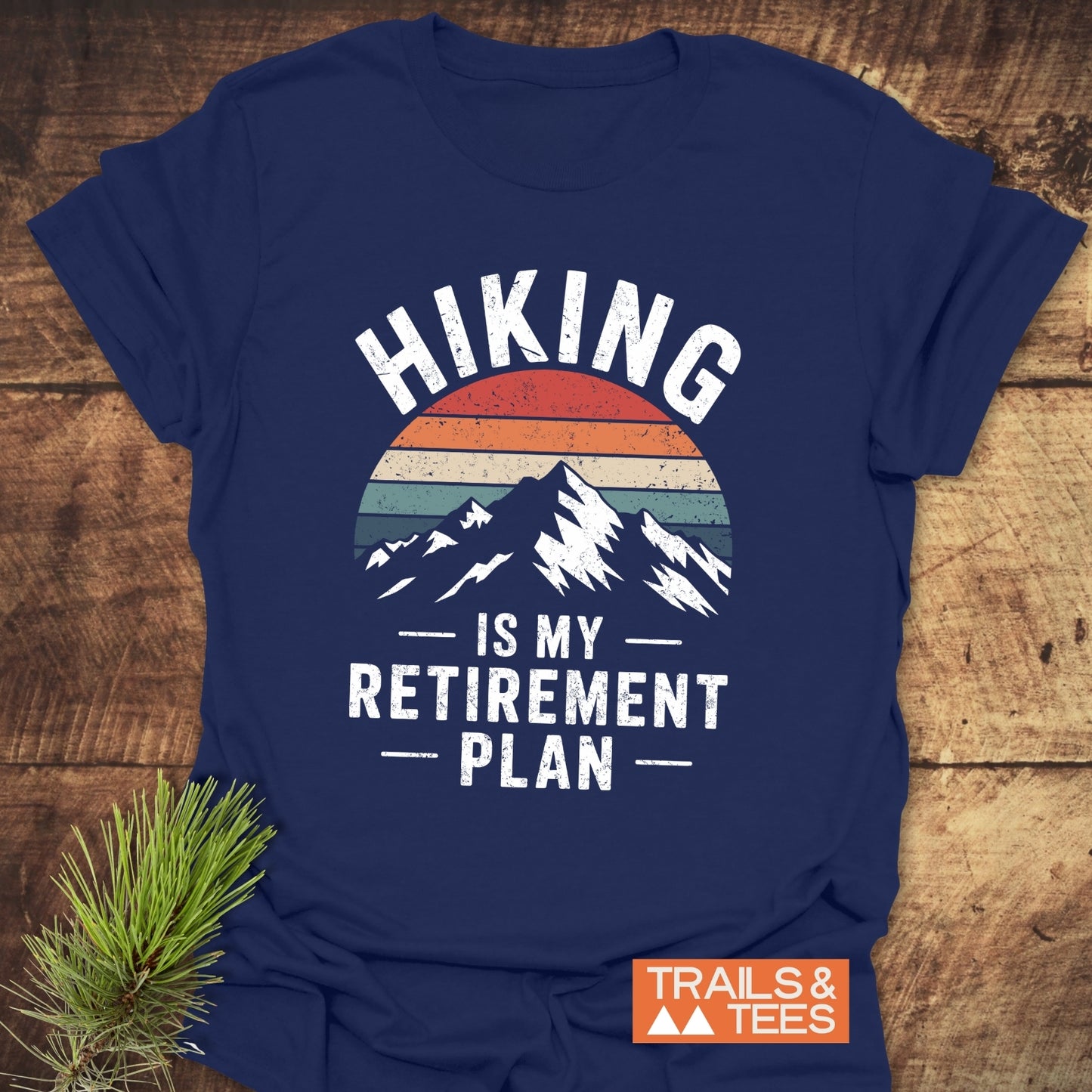 Navy-blue Hiking Is My Retirement Plan T-shirt with mountain graphic and retro stripes, featuring durable prints and soft cotton, laid flat on wood surface.