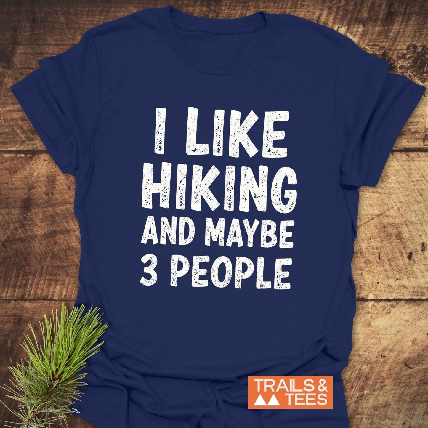 Navy-blue I Like Hiking And Maybe 3 People T-shirt, with distressed white slogan, orange TRAILS & TEES logo; laid on wood, pine sprig beside.