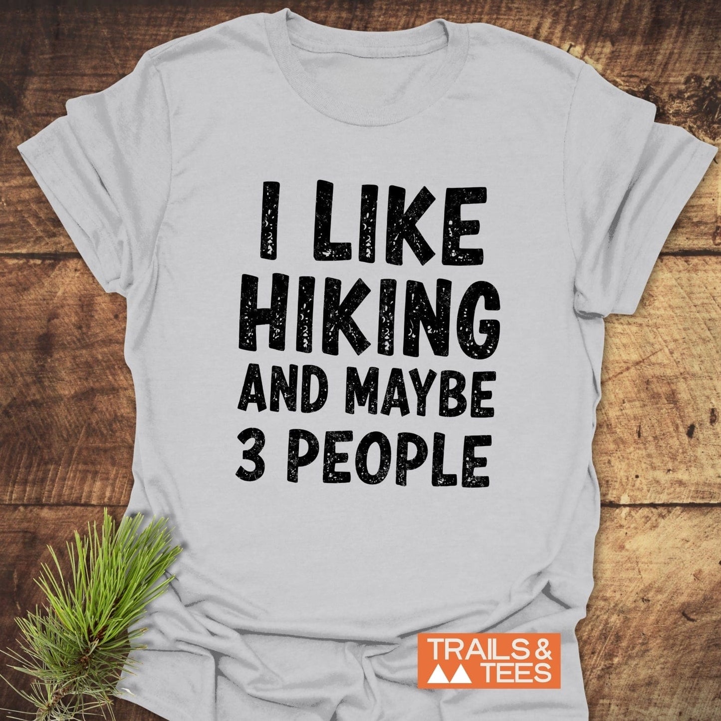 I Like Hiking And Maybe 3 People T-Shirt on wood, featuring distressed black text, pine sprig, and Trails & Tees logo.