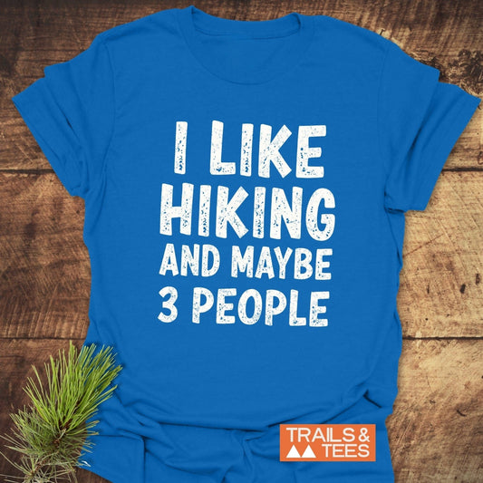 Bright blue I Like Hiking And Maybe 3 People T-shirt on rustic wood, featuring distressed white text and an orange TRAILS & TEES logo, with a pine sprig accent.