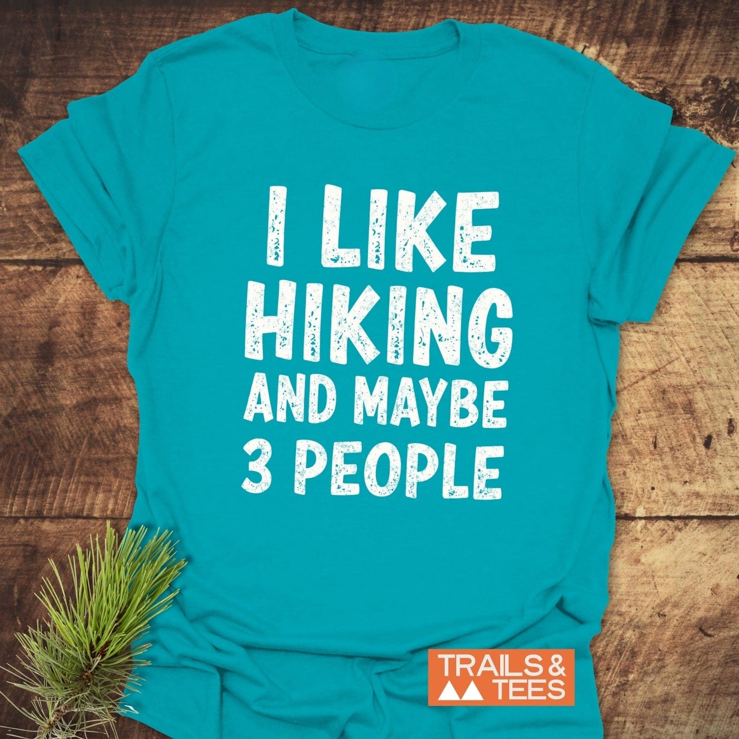 I Like Hiking And Maybe 3 People T-Shirt on wooden surface, showcasing bold white text and Trails & Tees logo, highlighting comfort and durability.