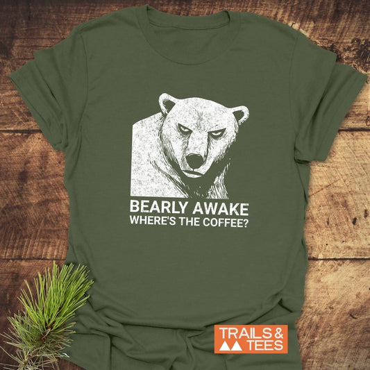 Bearly Awake T-Shirt featuring a distressed bear graphic and playful text, laid on wood with a pine branch, highlighting comfort and outdoor adventure spirit.