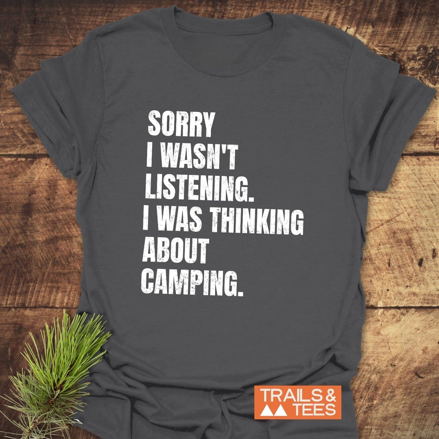 Sorry I Wasn't Listening Camping T-Shirt on wooden surface, featuring distressed white lettering and pine needle sprig. Made from soft cotton, ideal for outdoor enthusiasts.