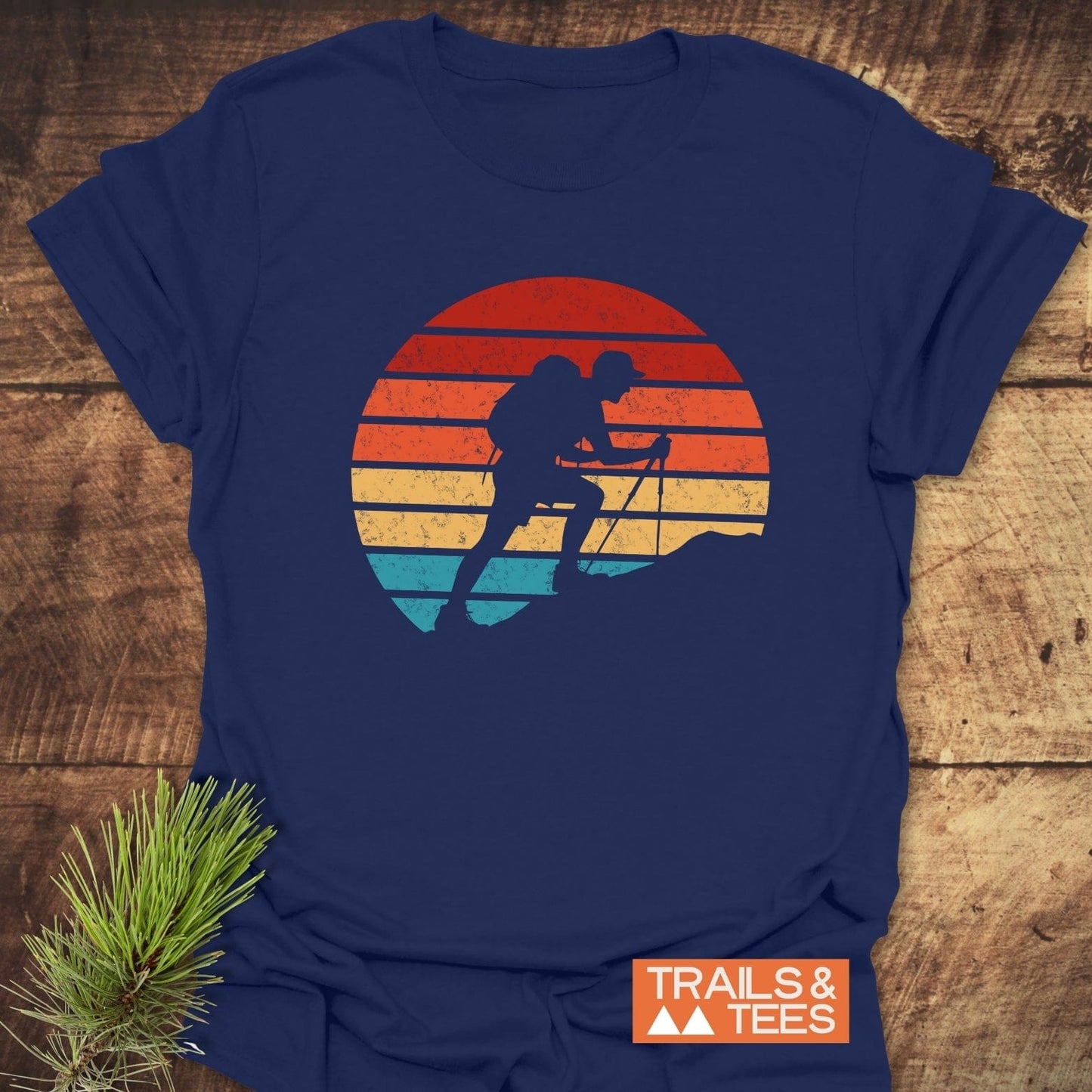 Uphill Hiker T-Shirt with vintage backpacker graphic, sunset motif. Laid flat on wood with pine sprig. Ideal for outdoor enthusiasts.