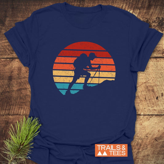 Uphill Hiker T-Shirt with vintage backpacker graphic, sunset motif. Laid flat on wood with pine sprig. Ideal for outdoor enthusiasts.