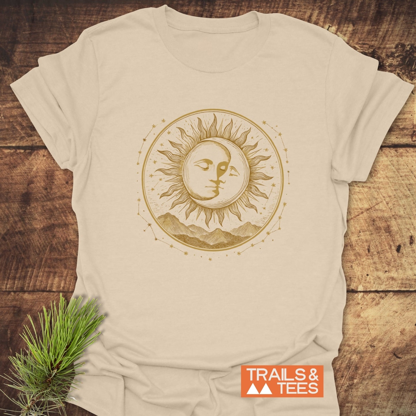 Celestial Embrace T-Shirt on wood, featuring a sun-moon face design with mountains, stars, and constellations; TRAILS & TEES logo at the bottom right.