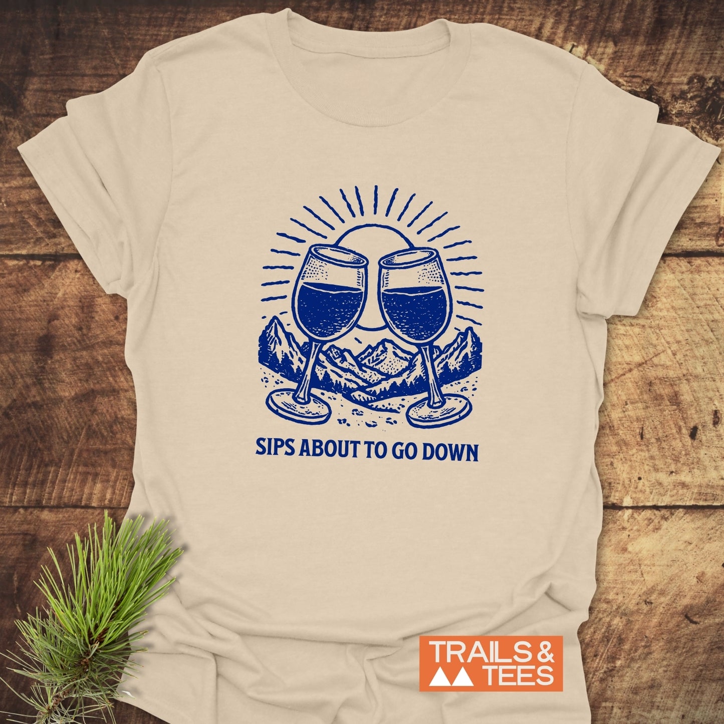 Light beige T-shirt featuring blue wine glasses, mountain, and sunburst design with SIPS ABOUT TO GO DOWN slogan; styled with evergreen sprig.