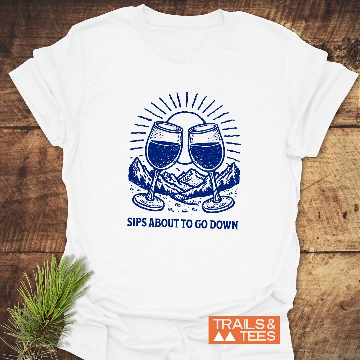 Sips About To Go Down T-Shirt showing two wine glasses and mountains, laid on wood, emphasizing soft cotton comfort and durability with the Trails & Tees logo.