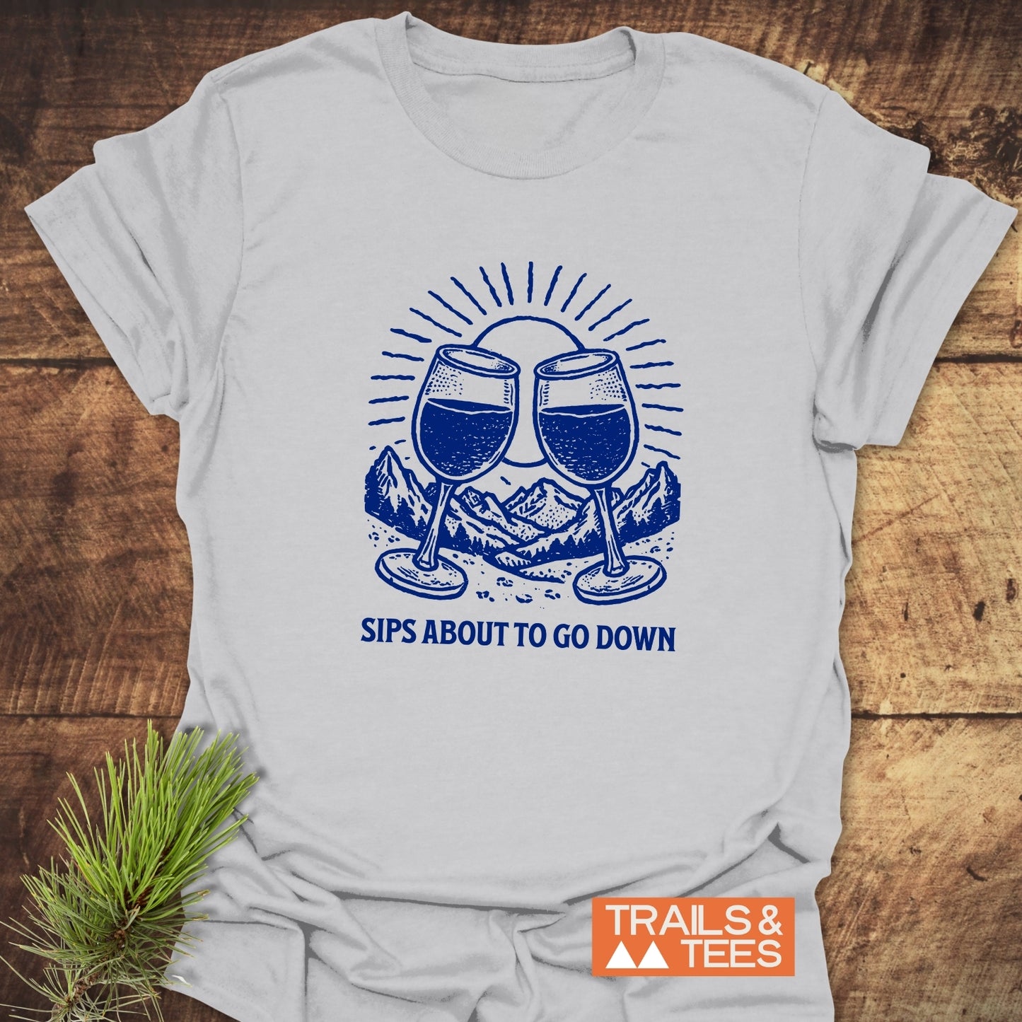 Sips About To Go Down T-Shirt featuring blue wine glass graphic on light fabric, laid on wood with pine bough; Trails & Tees logo visible.