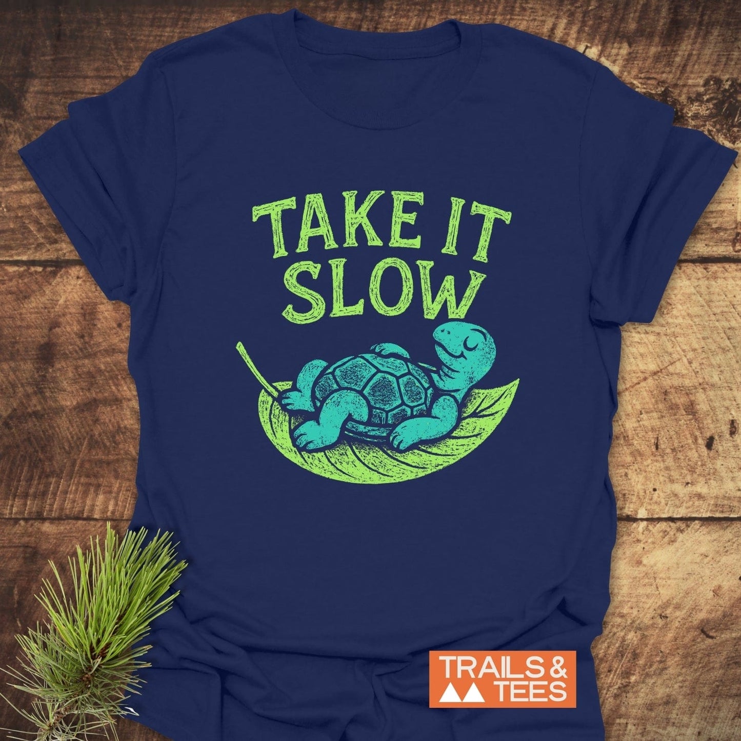 Navy-blue Take It Slow T-Shirt featuring a turtle graphic on a leaf hammock, emphasizing comfort and outdoor style. Includes TRAILS & TEES logo.