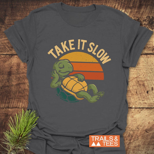 Take It Slow Turtle T-Shirt featuring a reclining cartoon turtle with a sunset backdrop and slogan, emphasizing comfort and durability for outdoor enthusiasts.