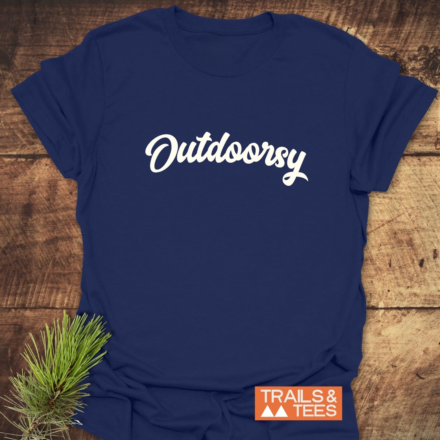 Outdoorsy T-Shirt with script font logo on chest, displayed on wood. Features a small TRAILS & TEES logo and a sprig of pine needles.