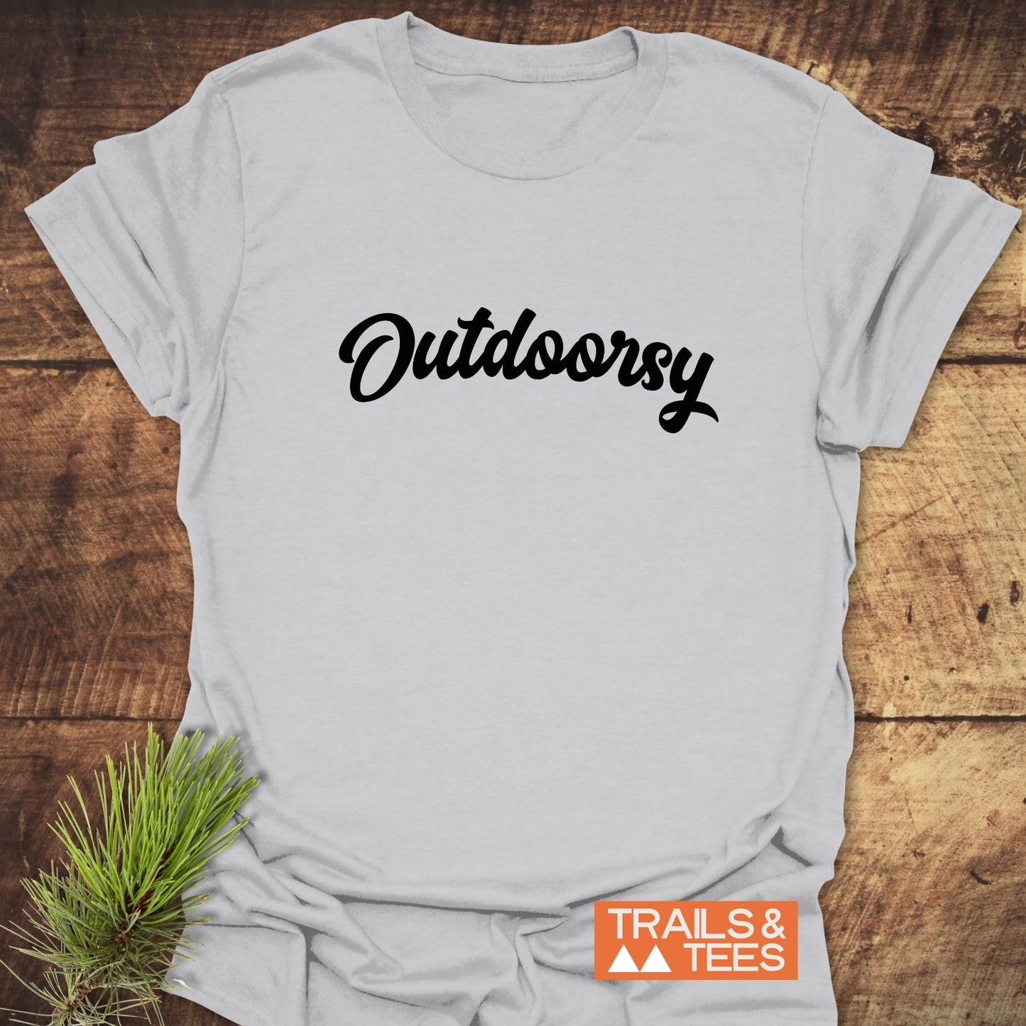 Outdoorsy T-Shirt with bold script on a rustic wooden surface, featuring a small TRAILS & TEES logo and a sprig of pine needles.