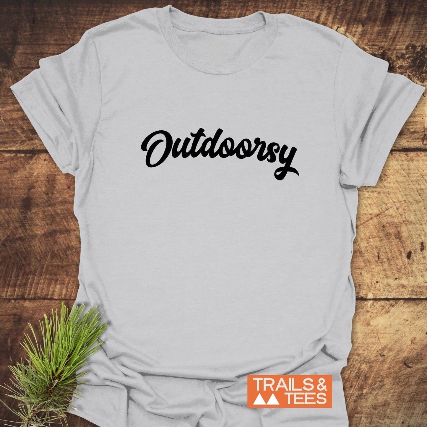Outdoorsy T-Shirt with bold script on a rustic wooden surface, featuring a small TRAILS & TEES logo and a sprig of pine needles.