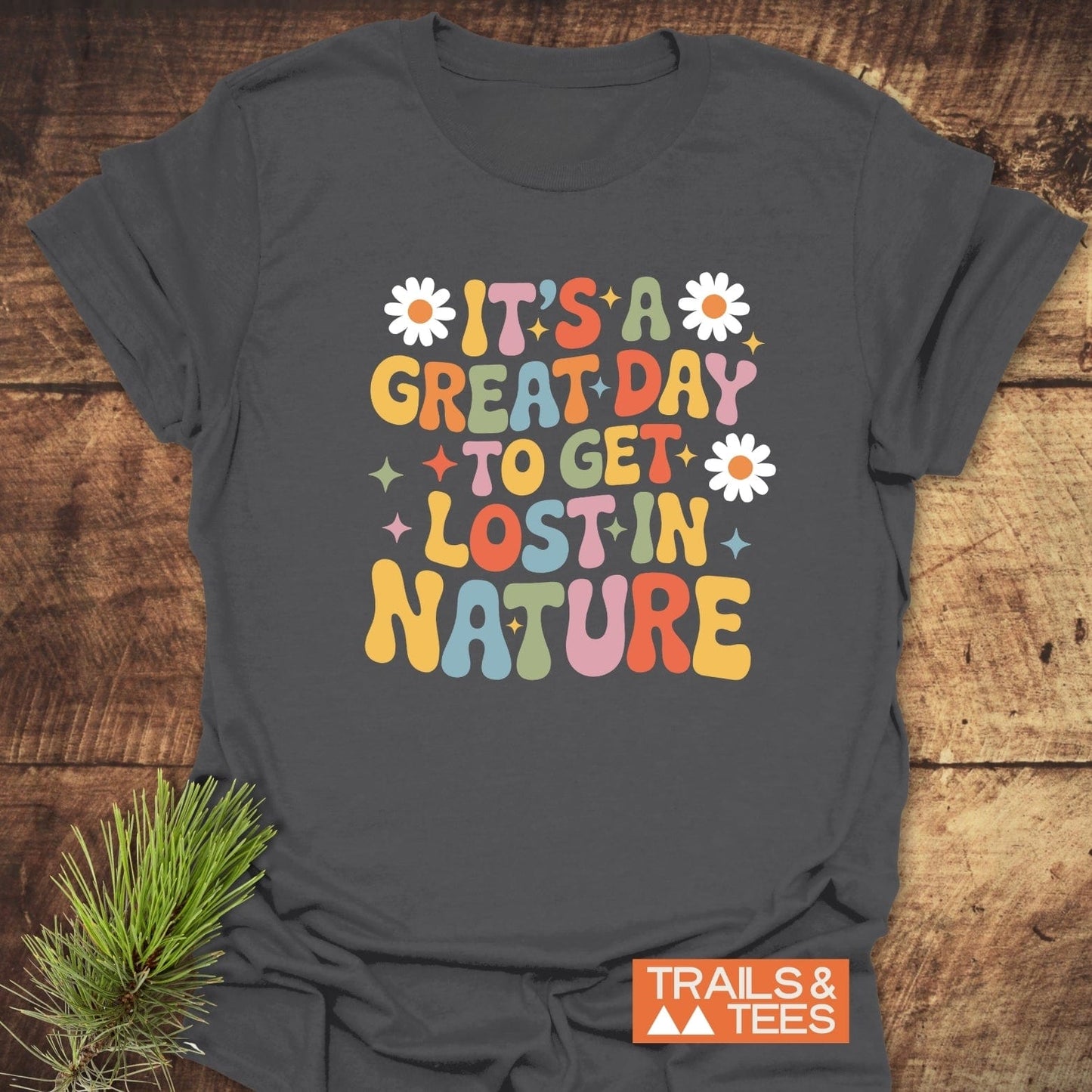 Dark-gray T-shirt with “IT’S A GREAT DAY TO GET LOST IN NATURE” slogan, daisy illustrations. Includes orange “TRAILS & TEES” logo, highlighting its outdoor adventure appeal.