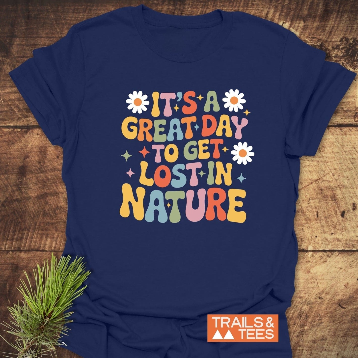 Navy-blue T-shirt with “It’s a great day to get lost in nature” slogan, featuring starbursts and daisies, next to a pine sprig and Trails & Tees logo.