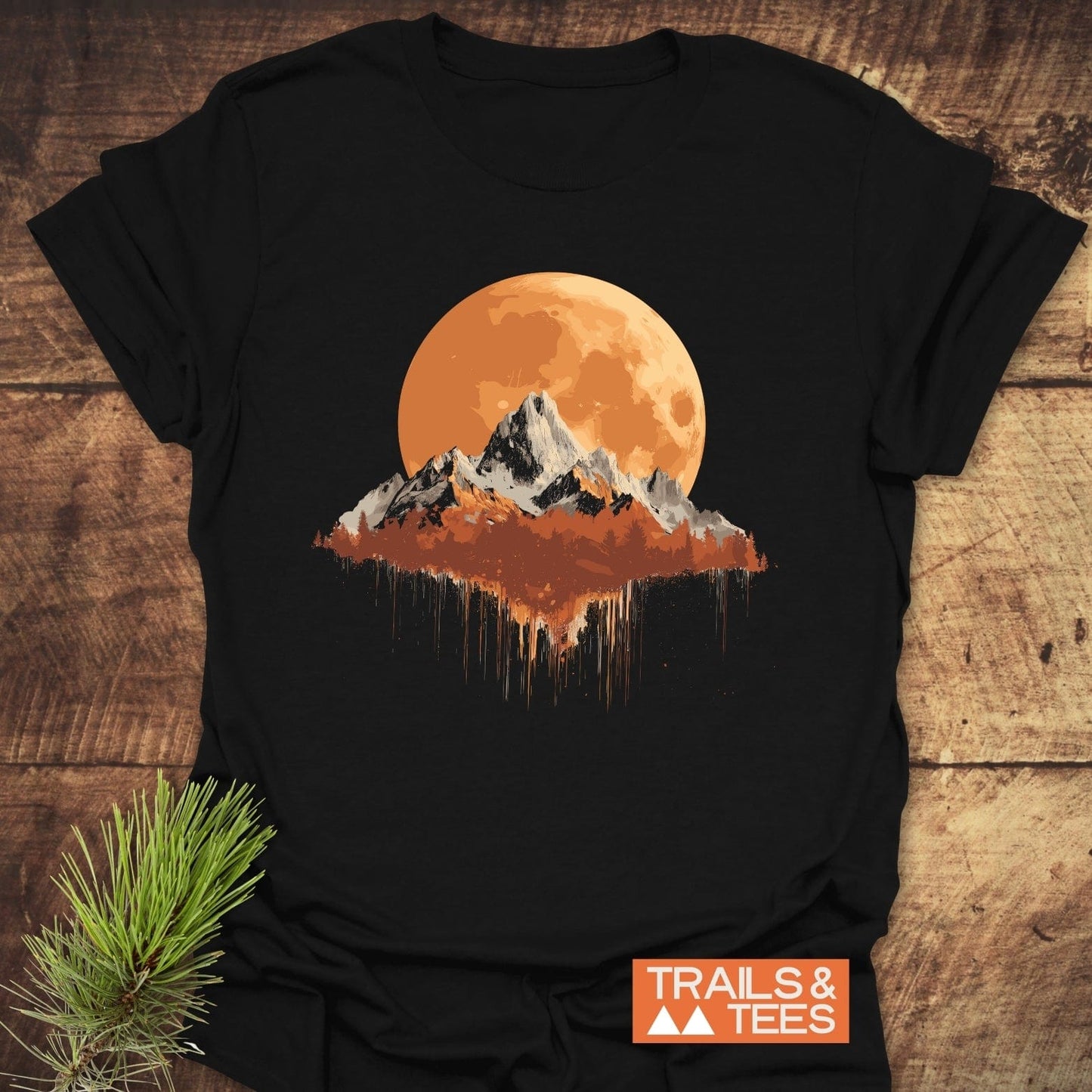 Orange Moon T-Shirt with a mountain range and paint-splatter effect on black fabric, enhanced by a rustic pine sprig and TRAILS & TEES logo.