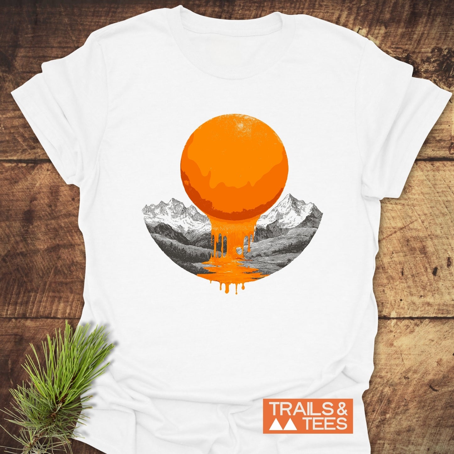 Melting Sun T-Shirt with a dripping sun graphic over mountains on white cotton, showcasing Trails & Tees' logo, laid flat on wood with pine sprig.