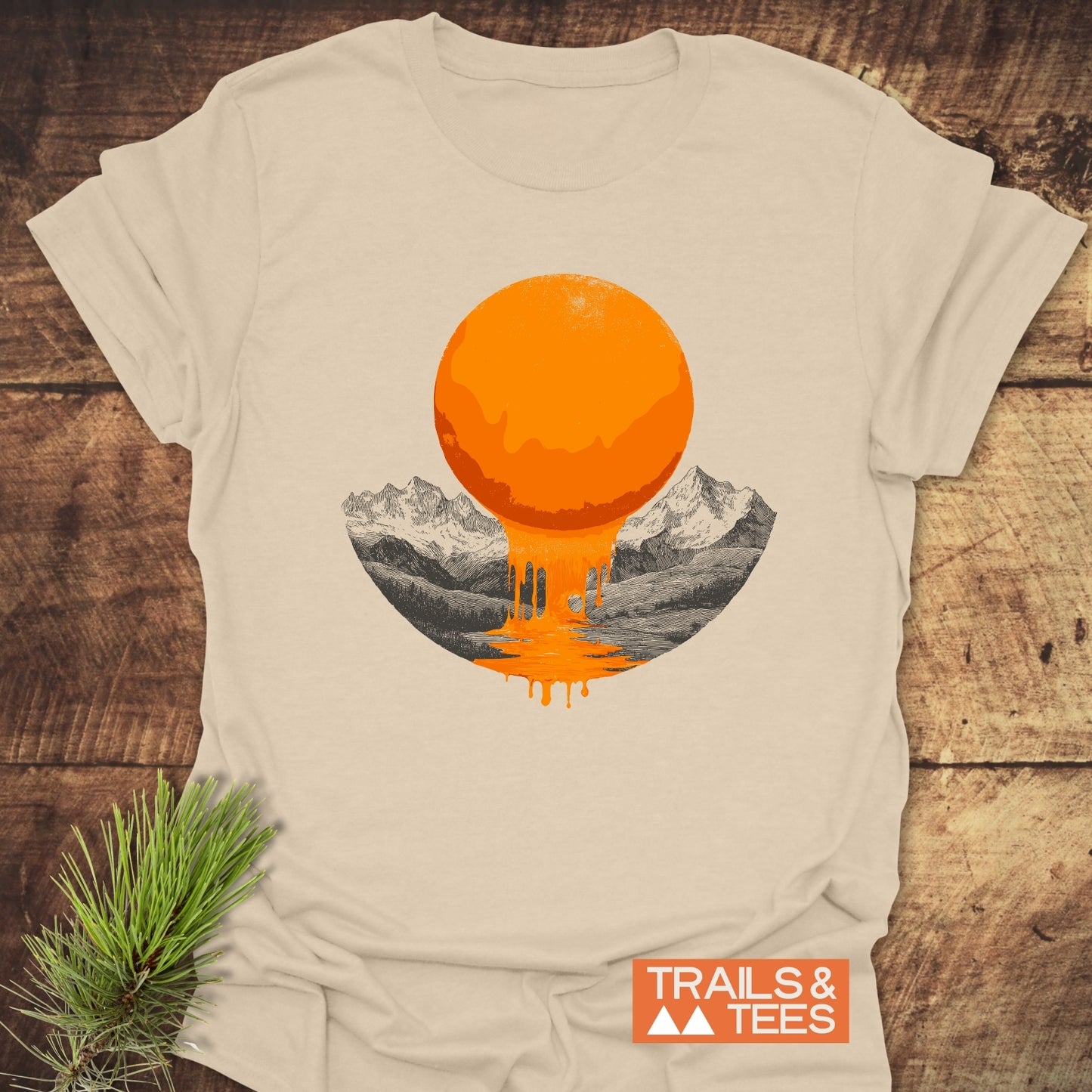 Melting Sun T-Shirt featuring a bright orange, dripping sun over sketched mountains; laid on a wooden surface with a pine sprig beside it.