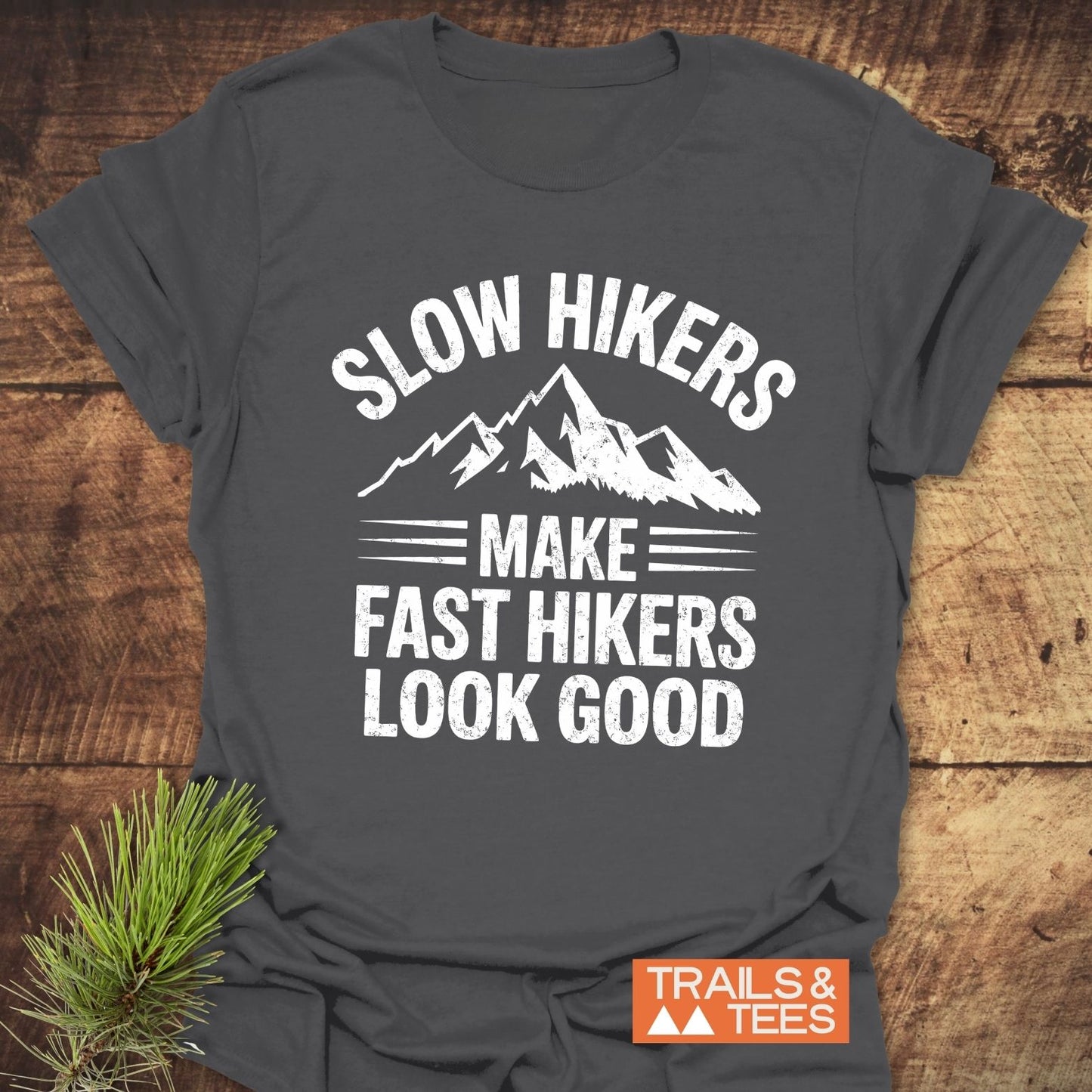 Dark-gray Slow Hikers T-Shirt with mountain graphic and slogan, featuring an orange TRAILS & TEES logo. Laid on wood with a pine sprig.