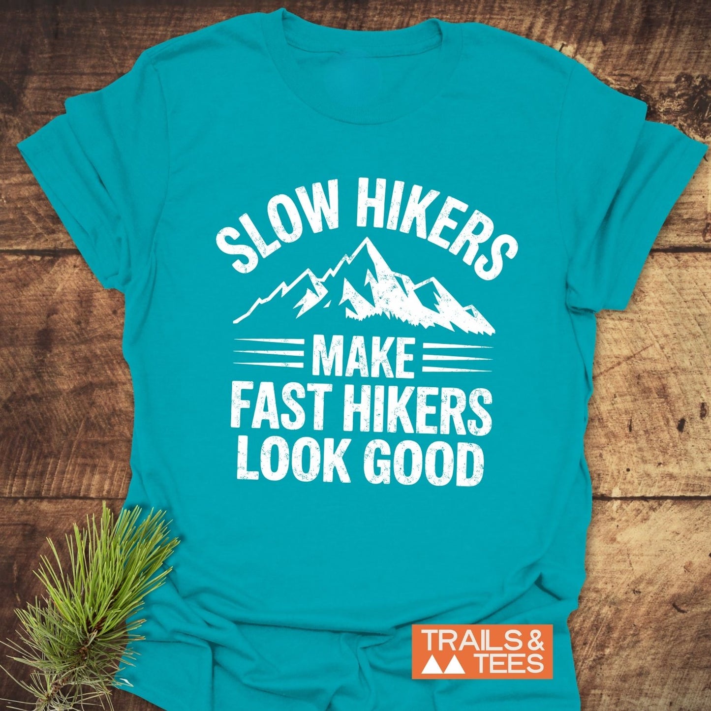 Slow Hikers T-Shirt in teal-blue featuring a mountain graphic and slogan, laid on wood. Soft, lightweight cotton, pre-shrunk for fit, with a durable print.