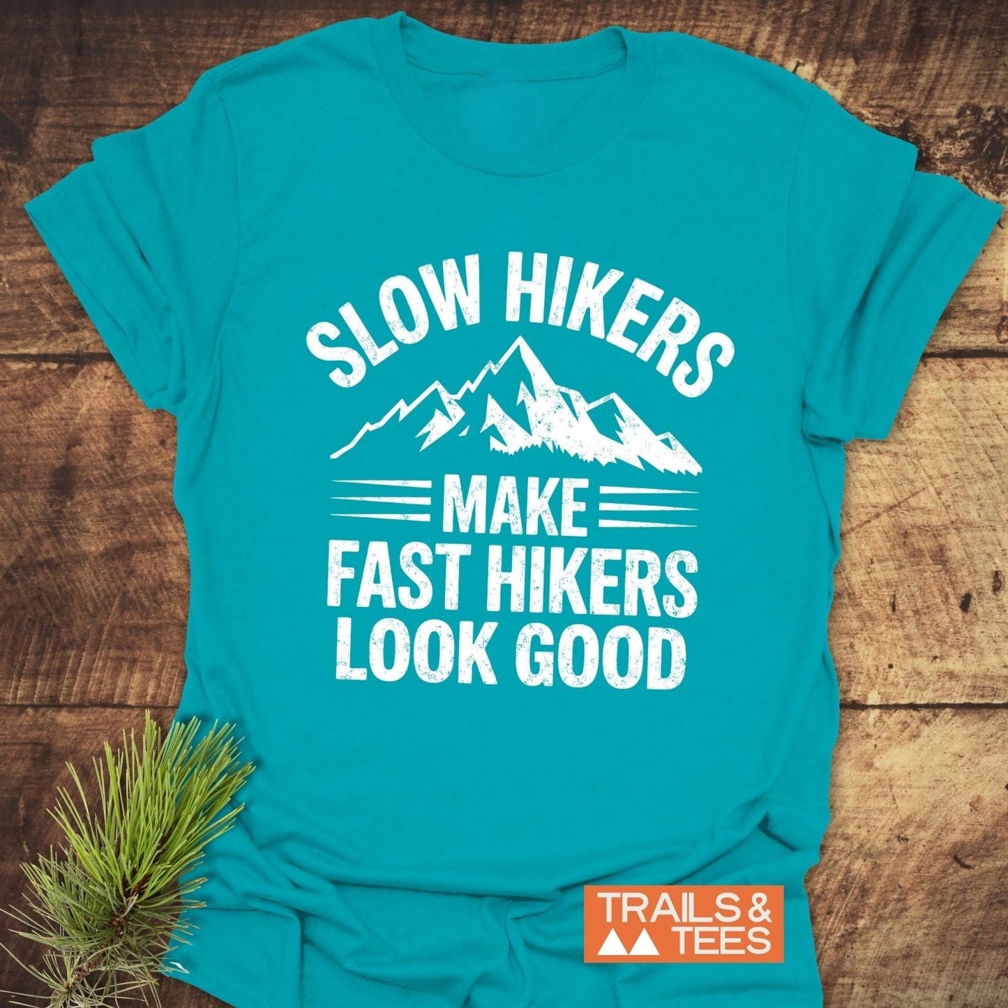Slow Hikers T-Shirt in teal-blue featuring a mountain graphic and slogan, laid on wood. Soft, lightweight cotton, pre-shrunk for fit, with a durable print.