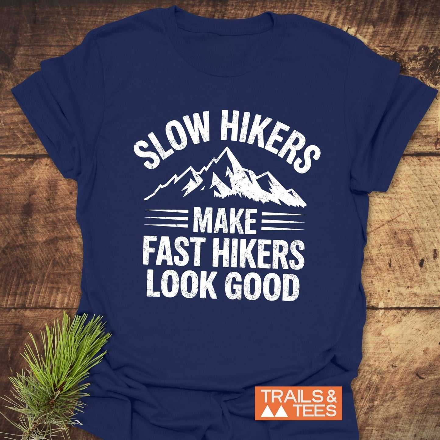 Navy-blue Slow Hikers T-Shirt with white mountain-themed text, laid on wood. Features orange label TRAILS & TEES and pine sprig beside it.