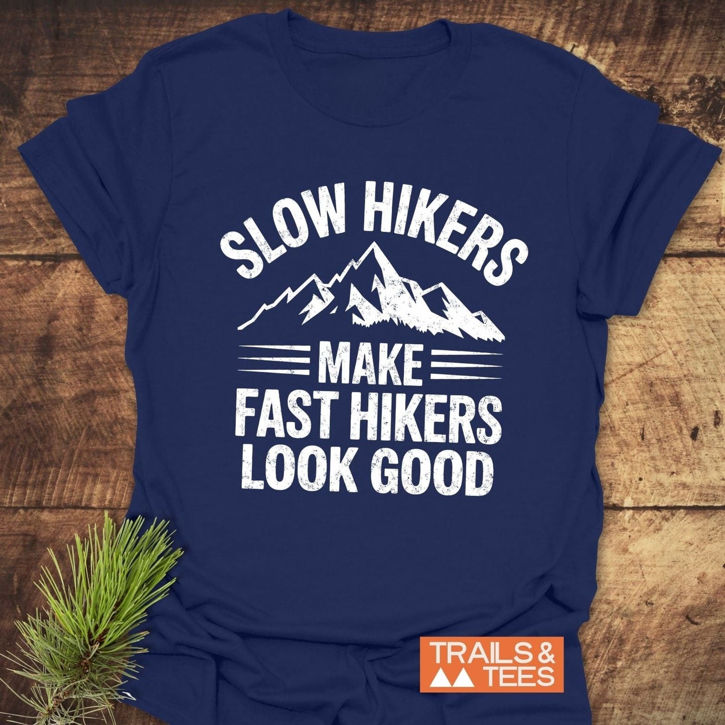 Navy-blue Slow Hikers T-Shirt with white mountain-themed text, laid on wood. Features orange label TRAILS & TEES and pine sprig beside it.