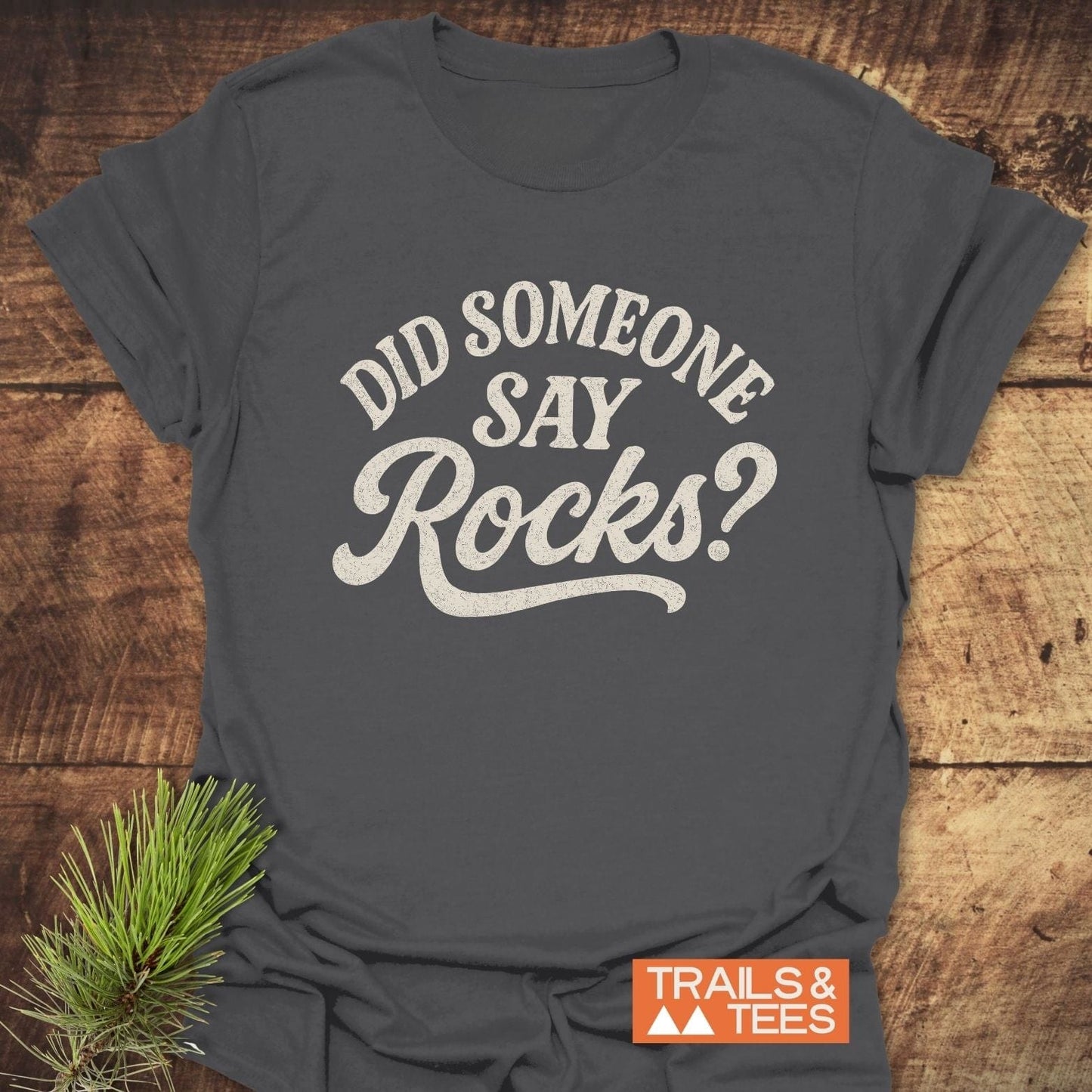 Did Someone Say Rocks? T-Shirt on wood, featuring distressed white script and Trails & Tees logo, with pine boughs. Soft cotton, durable print, pre-shrunk fit.