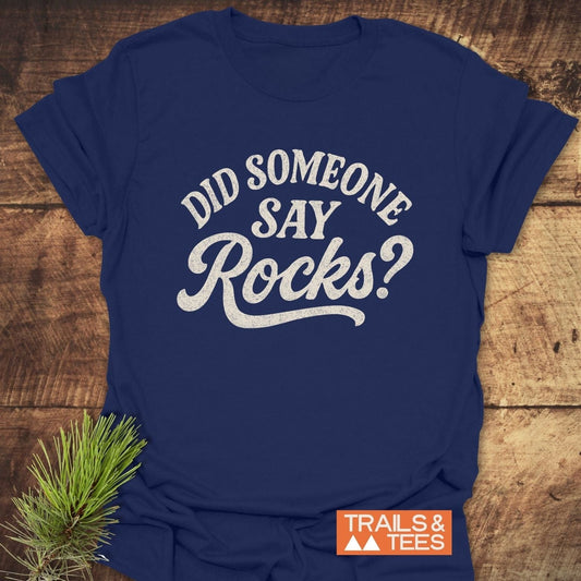 Navy-blue Did Someone Say Rocks? T-shirt with vintage white slogan, orange Trails & Tees logo, laid on wood with pine branch.