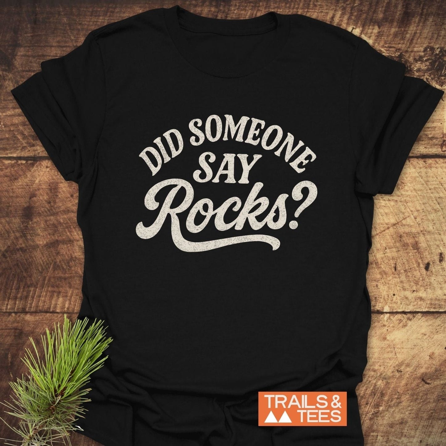 Flat-lay of 'Did Someone Say Rocks?' T-Shirt by TRAILS & TEES on wood, featuring distressed white lettering and rustic pine sprig accent.