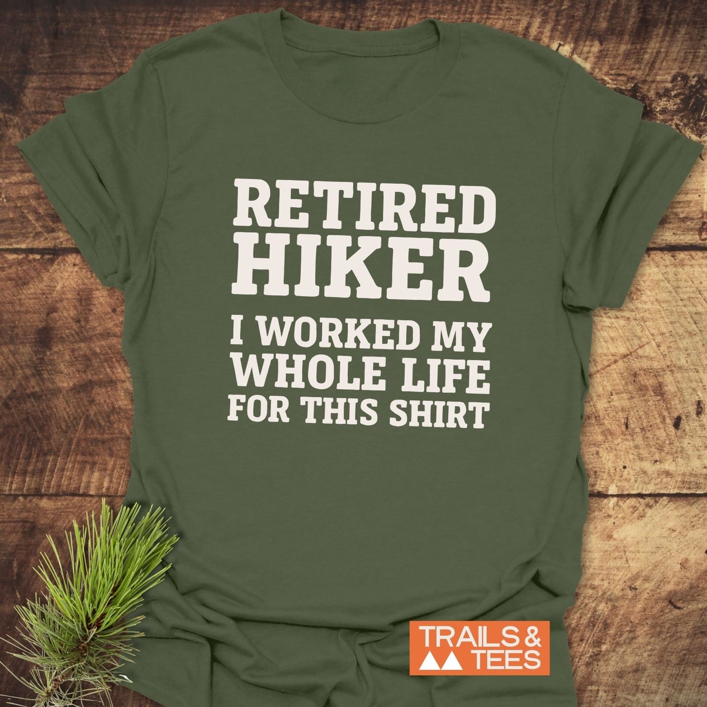 Army-green Retired Hiker T-shirt on wood surface, with bold white text. Features soft ring-spun cotton, durable print, and TRAILS & TEES logo.