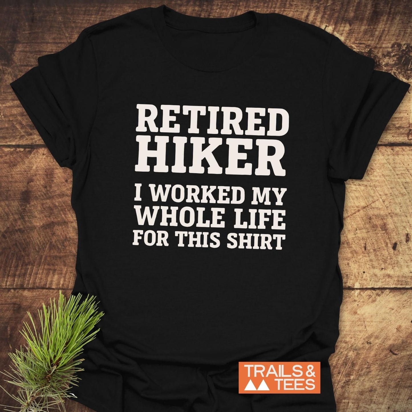Black Retired Hiker T-shirt with white text, laid flat. Includes TRAILS & TEES logo and pine branch for outdoor-themed staging.
