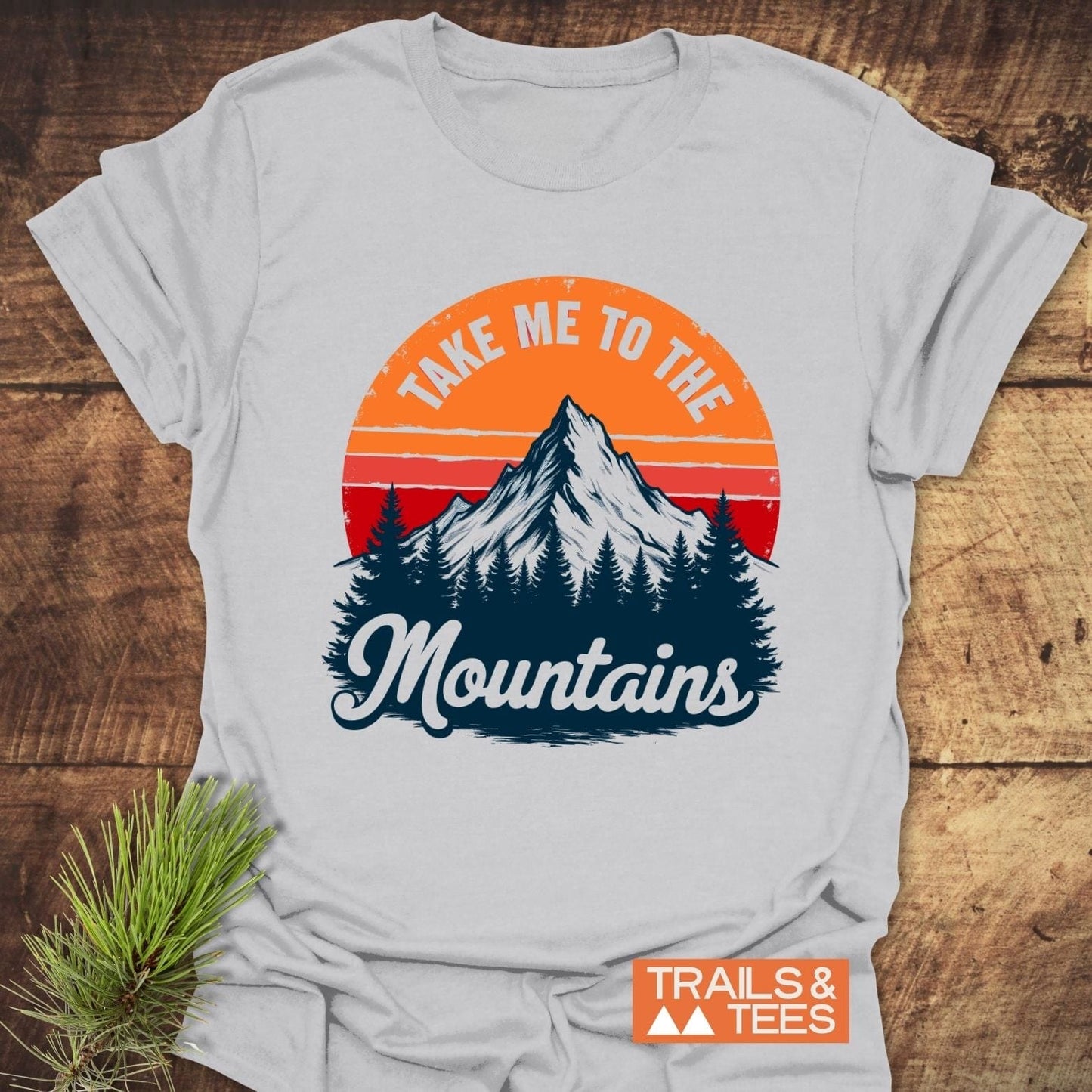 Take Me To The Mountains T-Shirt featuring a stylized mountain scene with pine trees and sunset, crafted from soft cotton for comfort and durability.