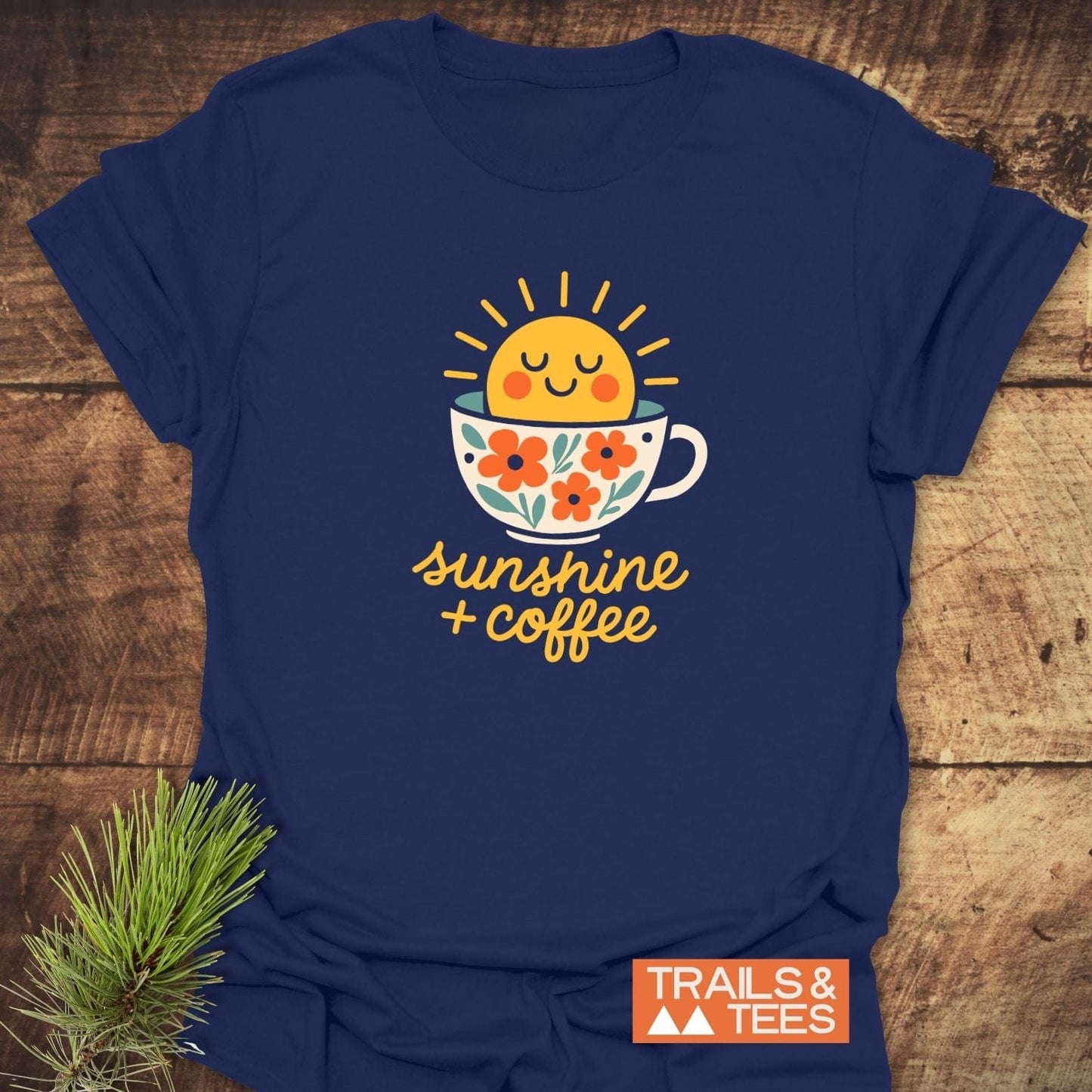 Sunshine And Coffee T-Shirt with a smiling sun in a coffee cup graphic, crafted from soft cotton, featuring sunshine + coffee text, and a Trails & Tees logo.