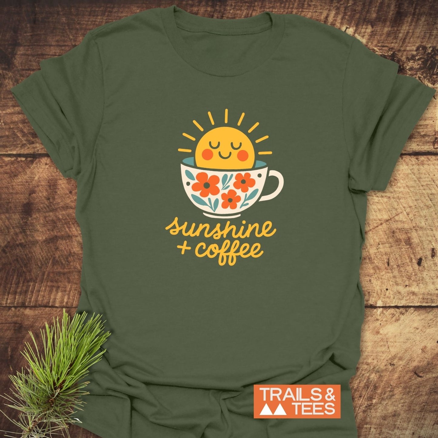 Sunshine And Coffee T-Shirt featuring a sun in a coffee cup with floral accents, laid flat on wood, embodying outdoor adventure and comfort by TRAILS & TEES.