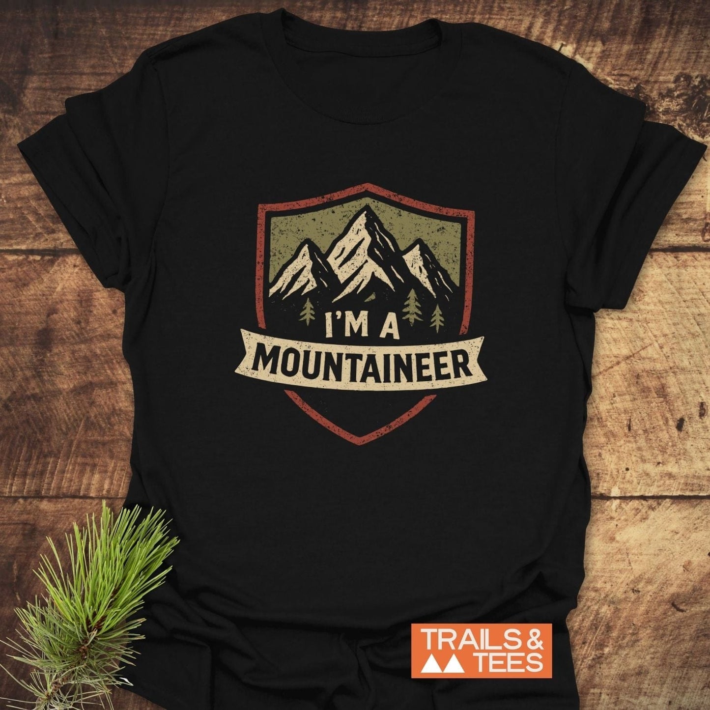 Mountaineer T-Shirt featuring a mountain-shield graphic with I'm a Mountaineer banner, laid on wood. Includes TRAILS & TEES label and pine sprig accent.